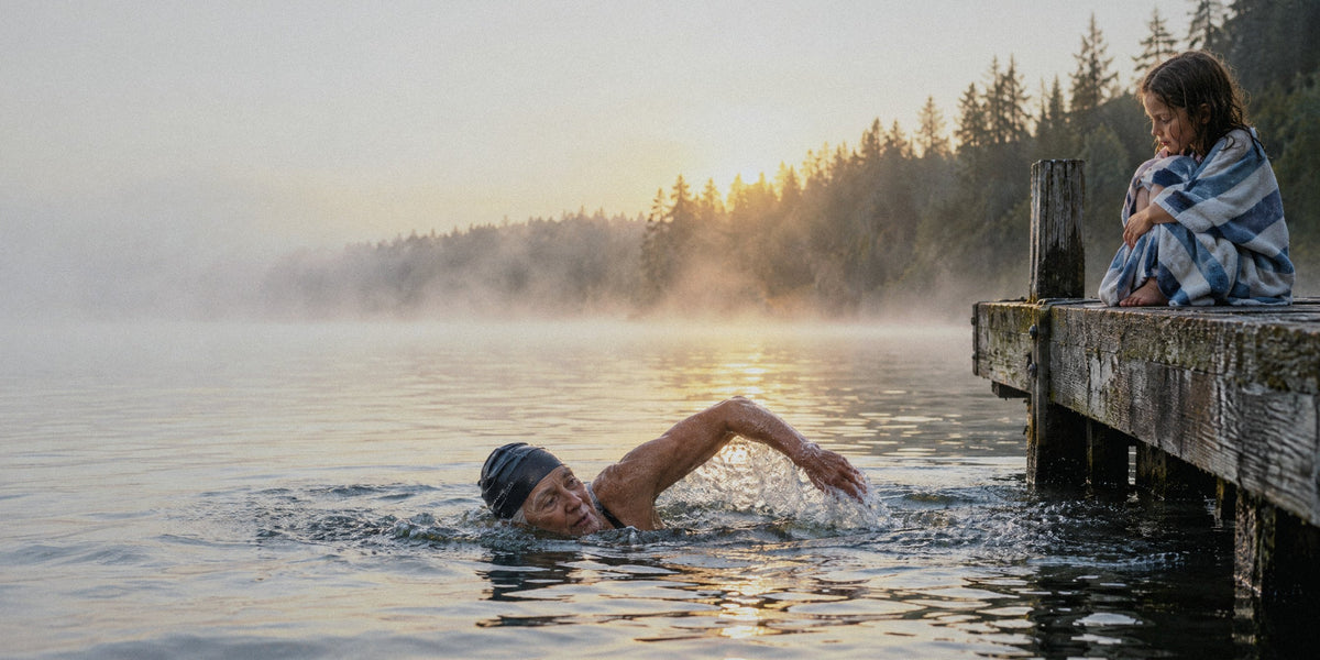 A grandmother swimming in a misty lake at sunrise while her granddaughter watches from the dock